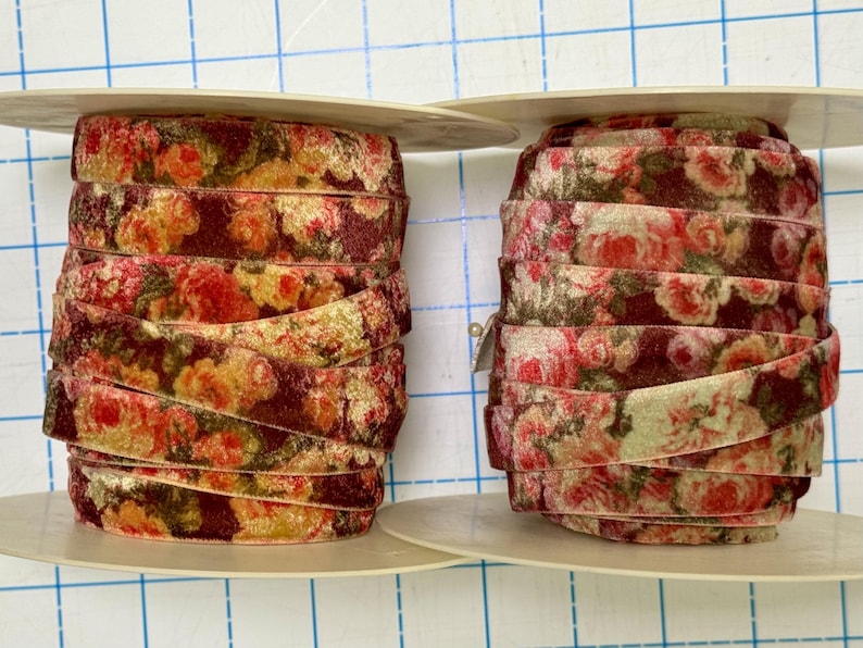 May include: Two spools of velvet ribbon featuring a floral pattern with shades of deep red, burgundy, and muted yellow. The ribbon is wound tightly around circular plastic spools, displaying a repeating rose print across the soft fabric surface.