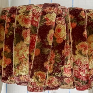 May include: A spool of velvet ribbon featuring a deep burgundy background with a repeating floral pattern in shades of cream, muted orange, and dark green. The ribbon is wound tightly, displaying the soft, textured surface of the fabric.