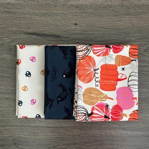 May include: Three folded pieces of fabric. The first is cream with small orange, pink, and black skull icons. The second is dark navy blue with black bird silhouettes. The third is white with a pattern of orange, pink, and black pumpkins and spiders.