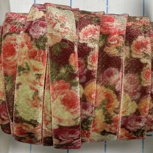 May include: A spool of velvet ribbon featuring a floral pattern with shades of deep red, soft pink, and muted green. The fabric has a textured, soft surface with a subtle sheen, wound tightly to display the continuous rose-themed print.