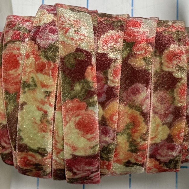 May include: A spool of velvet ribbon featuring a floral pattern with shades of deep red, soft pink, and muted green. The fabric has a textured, soft surface with a subtle sheen, wound tightly to display the continuous rose-themed print.