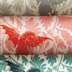 May include: Three rolls of fabric featuring a repeating pattern of winged dragons and leafy foliage. The top roll is grey, the middle roll is pink, and the bottom roll is teal. Each dragon and leaf is outlined with a metallic gold ink.
