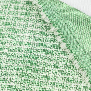 May include: A piece of green and white woven fabric with a textured, grid-like pattern. The material features a folded corner revealing a frayed, raw edge with visible white threads along the seam, showcasing the dense, interlaced weave structure.
