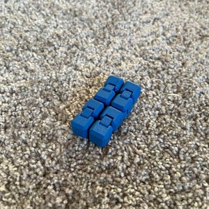 Può includere: Un giocattolo fidget blu composto da blocchi rettangolari collegati. Il giocattolo è di un blu vibrante e si trova su un tappeto grigio strutturato. I blocchi sono progettati per essere manipolati e spostati.
