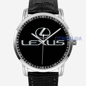 May include: A wristwatch with a silver-tone circular case and a black dial featuring the silver Lexus logo and text. The watch has silver hour, minute, and second hands, a black leather strap with a textured finish, and a crown on the right side.