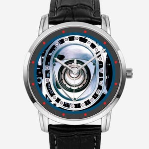 Pode incluir: Um relógio de pulso com caixa circular prateada e pulseira de couro preta. O mostrador apresenta um gráfico detalhado de um rotor de motor rotativo, rodeado por um anel azul escuro com pequenos pontos vermelhos marcando as horas e ponteiros prateados.