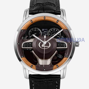 Pode incluir: Um relógio de pulso com caixa de metal prateado e bracelete de couro preto texturizado. O mostrador apresenta uma impressão gráfica de um volante e painel de carro com um logótipo Lexus ao centro. Três ponteiros prateados.