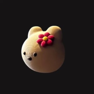 May include: A light beige, bear-shaped decorative item with a red and yellow flower on its head. The bear has small, black eyes and a textured surface, set against a dark background.