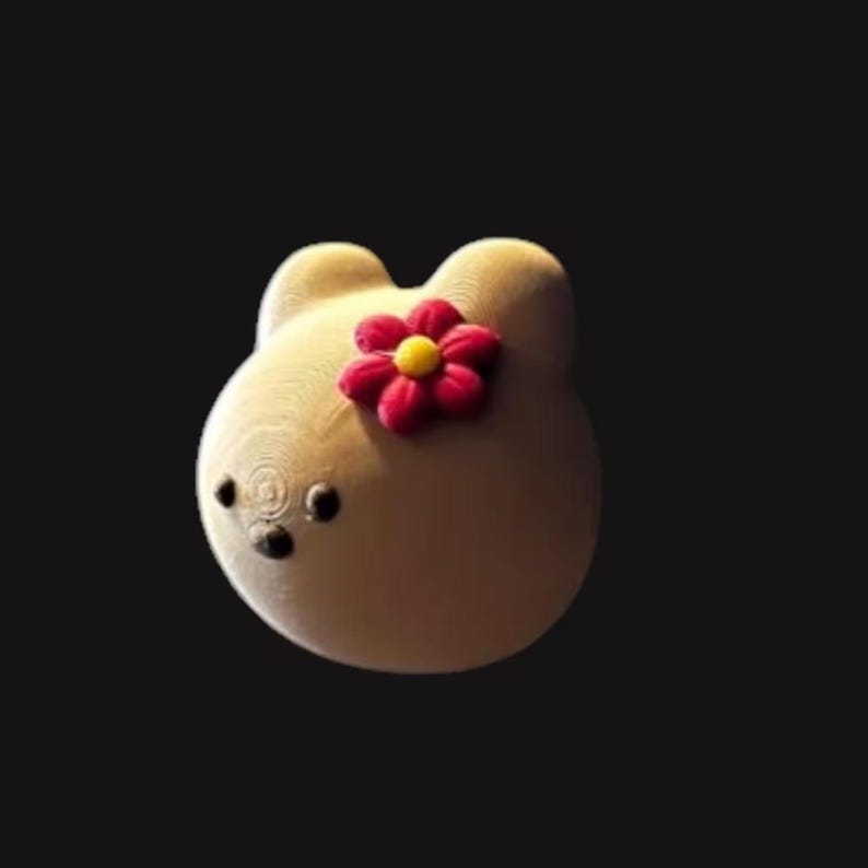 May include: A light beige, bear-shaped decorative item with a red and yellow flower on its head. The bear has small, black eyes and a textured surface, set against a dark background.