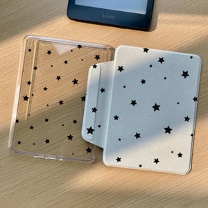 May include: A Kindle e-reader with a white cover featuring a black star pattern, and a clear protective case with black star accents. The e-reader is black, and the cases are open on a wooden surface.