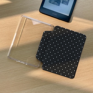 May include: A clear protective case for an e-reader and a black cover featuring a white polka dot pattern. The black cover is rectangular with rounded corners and is positioned partially inside the clear case, alongside a black e-reader device.