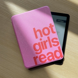 Puede incluir: Una funda para lector electrónico de color rosa con el texto "hot girls read" en letras rojas en negrita. La funda está colocada sobre un lector electrónico negro con pantalla visible, mostrando una superficie texturizada y forma rectangular.