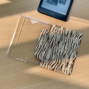 May include: A Kindle e-reader with a clear protective case and a patterned cover featuring a cream and brown zebra stripe design. The e-reader is black and rests on a light wood surface.