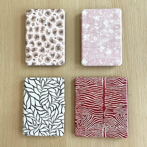 May include: Four rectangular e-reader cases arranged in a grid. The top left case features a tan and brown leopard print. The top right case has a dusty pink background with white floral branches. The bottom left case shows a black and white leaf pattern. The bottom right case displays a deep red and white zebra stripe design.