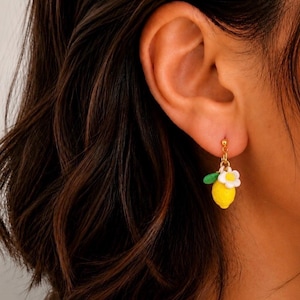 Puede incluir: Un pendiente dorado con un dije de limón amarillo, una flor blanca y una hoja verde. El pendiente se lleva en la oreja de una persona con cabello castaño oscuro.