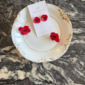 Può includere: Tre orecchini a lobo a forma di papavero rosso brillante con centri neri. Due orecchini sono fissati su un cartoncino rettangolare bianco con la scritta FRANEVA ATELIER HANDMADE JEWELRY, mentre il terzo orecchino poggia su un piattino bianco.