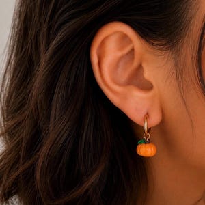 Può includere: Un orecchino a cerchio dorato con un piccolo ciondolo a forma di zucca arancione, caratterizzato da un minuscolo dettaglio a foglia verde. La zucca è sospesa a un sottile cerchio in metallo dorato che attraversa il lobo dell'orecchio.
