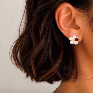 Könnte beinhalten: Weiße Stecker-Ohrringe in Form einer Gänseblümchenblüte mit gelbem Zentrum und rotem Akzent. Die Ohrringe werden am Ohr einer Person getragen, mit dunkelbraunem Haar im Hintergrund. Die Ohrringe sind ein Blumendesign.