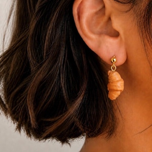 Può includere: Orecchino a lobo dorato con una piccola perla rotonda e un ciondolo a forma di croissant marrone chiaro. L'orecchino è indossato su un orecchio, con capelli castano scuro sullo sfondo.