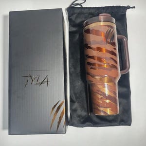 May include: A brown and copper-colored travel mug with a handle and a lid, featuring a tiger stripe pattern. The mug is next to a black box with the text "TMA" and a claw mark design. A black drawstring bag is also included.