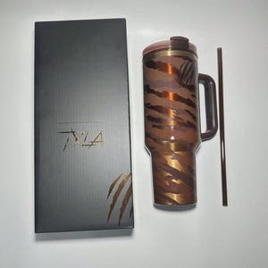 May include: A brown and gold tumbler with a handle and a matching straw, next to a black box with the text "TYLA". The tumbler has a tiger stripe pattern. The box has a palm leaf design.