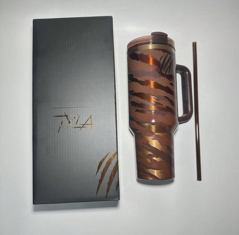 May include: A brown and gold tumbler with a handle and a matching straw, next to a black box with the text "TYLA". The tumbler has a tiger stripe pattern. The box has a palm leaf design.