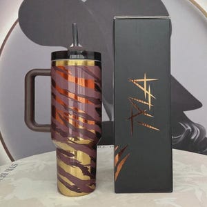 May include: A stainless steel tumbler with a handle, straw, and lid. The tumbler has a brown, gold, and orange tiger stripe pattern. A black box with gold text is next to the tumbler.