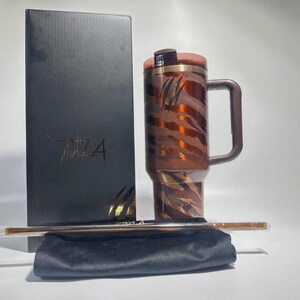 May include: A brown and copper-colored travel mug with a handle, featuring a tiger stripe design and the word "TIZA" on the box. Includes a straw and a black carrying pouch.