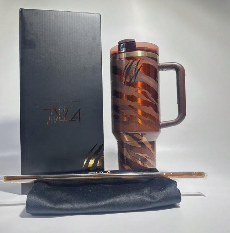 May include: A brown and copper-colored travel mug with a handle, featuring a tiger stripe design and the word "TIZA" on the box. Includes a straw and a black carrying pouch.