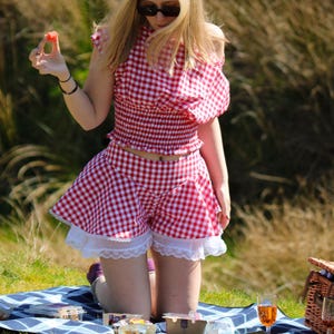May include: A red and white gingham two-piece outfit, comprising a crop top and shorts, worn with white lace trim. The outfit is part of a picnic scene, with a picnic basket and blanket visible.