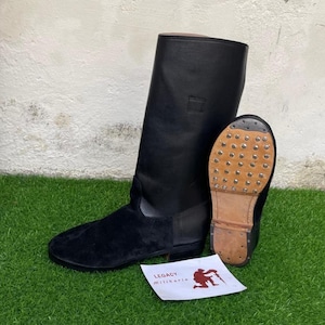 May include: Black leather military boot with a suede vamp and a studded sole. The boot is tall, with a smooth leather upper and a small square patch near the top. A second boot is shown with the sole facing up. A small paper with the text "LEGACY Militaria" is in the image.