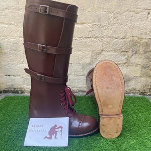 Puede incluir: Un par de botas altas de cuero marrón con múltiples correas con hebillas y cordones rojos. Una bota está de pie y la otra está tumbada de lado, mostrando la suela. Una tarjeta con el texto "LEGACY Militaria" está delante de las botas.