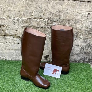 Peut inclure: Deux bottes d'équitation en cuir marron, hautes jusqu'aux genoux, avec une finition lisse et un bout carré. Une carte blanche avec le texte "LEGACY Militaria" et une silhouette d'une personne est sur l'herbe.