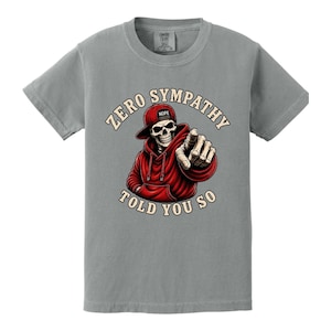 May include: Gray t-shirt featuring a graphic of a skeleton wearing a red hoodie and hat, pointing. The text reads "ZERO SYMPATHY TOLD YOU SO". The hat has the word "NOPE" on it.