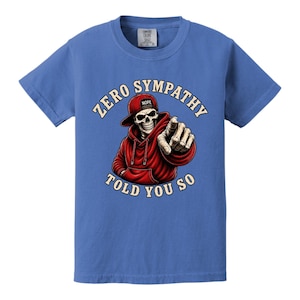 May include: Blue t-shirt featuring a graphic of a skeleton wearing a red hoodie and hat, pointing. The text reads "ZERO SYMPATHY TOLD YOU SO". The hat has the word "NOPE" on it.