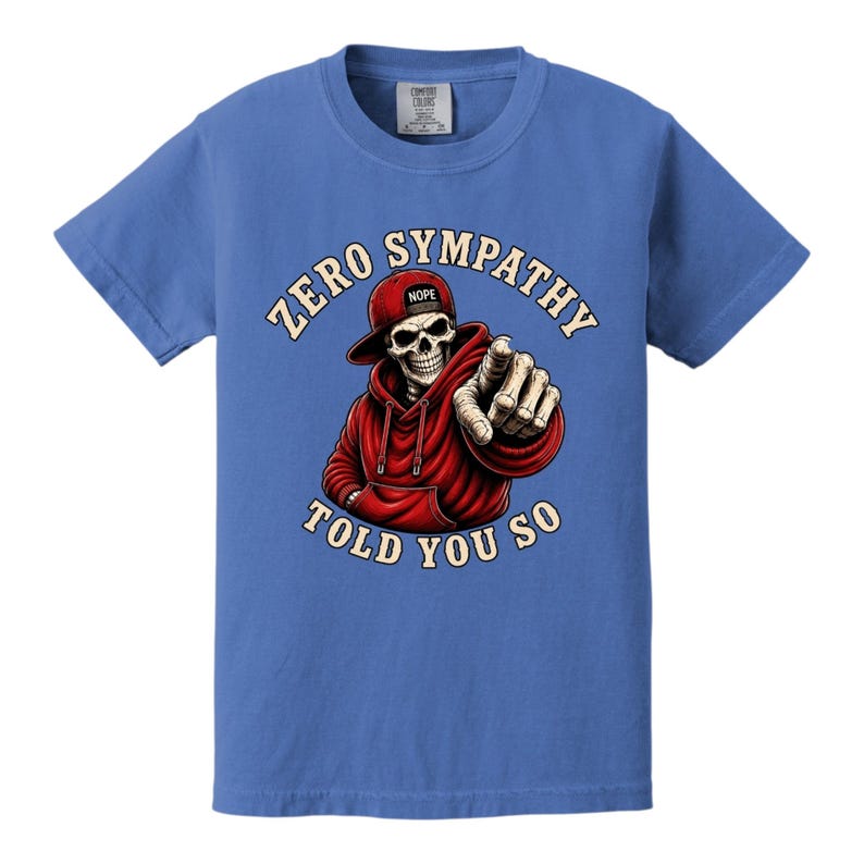 May include: Blue t-shirt featuring a graphic of a skeleton wearing a red hoodie and hat, pointing. The text reads "ZERO SYMPATHY TOLD YOU SO". The hat has the word "NOPE" on it.
