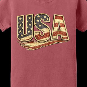 May include: A maroon-colored t-shirt featuring the word "USA" in a distressed, vintage style. The letters are designed with the American flag, including stars and stripes, and a gold outline. The shirt is a Comfort Colors brand.