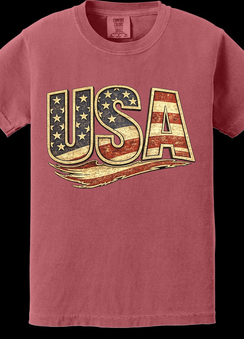 May include: A maroon-colored t-shirt featuring the word "USA" in a distressed, vintage style. The letters are designed with the American flag, including stars and stripes, and a gold outline. The shirt is a Comfort Colors brand.