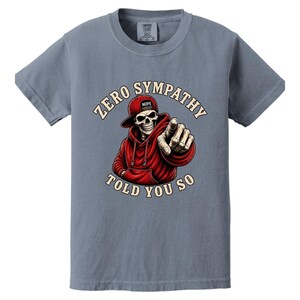 May include: A blue-grey t-shirt featuring a graphic of a skeleton wearing a red hoodie and hat, pointing at the viewer. The text "ZERO SYMPATHY TOLD YOU SO" is arched around the skeleton. The hat has the word "NOPE" on it.