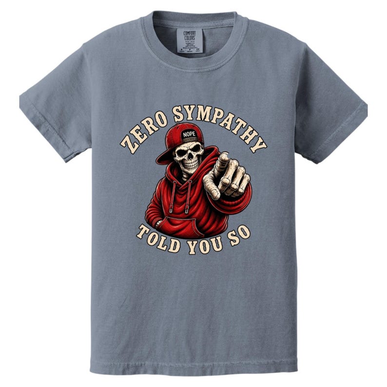 May include: A blue-grey t-shirt featuring a graphic of a skeleton wearing a red hoodie and hat, pointing at the viewer. The text "ZERO SYMPATHY TOLD YOU SO" is arched around the skeleton. The hat has the word "NOPE" on it.