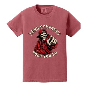 May include: A maroon t-shirt featuring a graphic of a skeleton wearing a red hoodie and hat, pointing. The text reads "ZERO SYMPATHY TOLD YOU SO". The hat has the word "NOPE" on it.