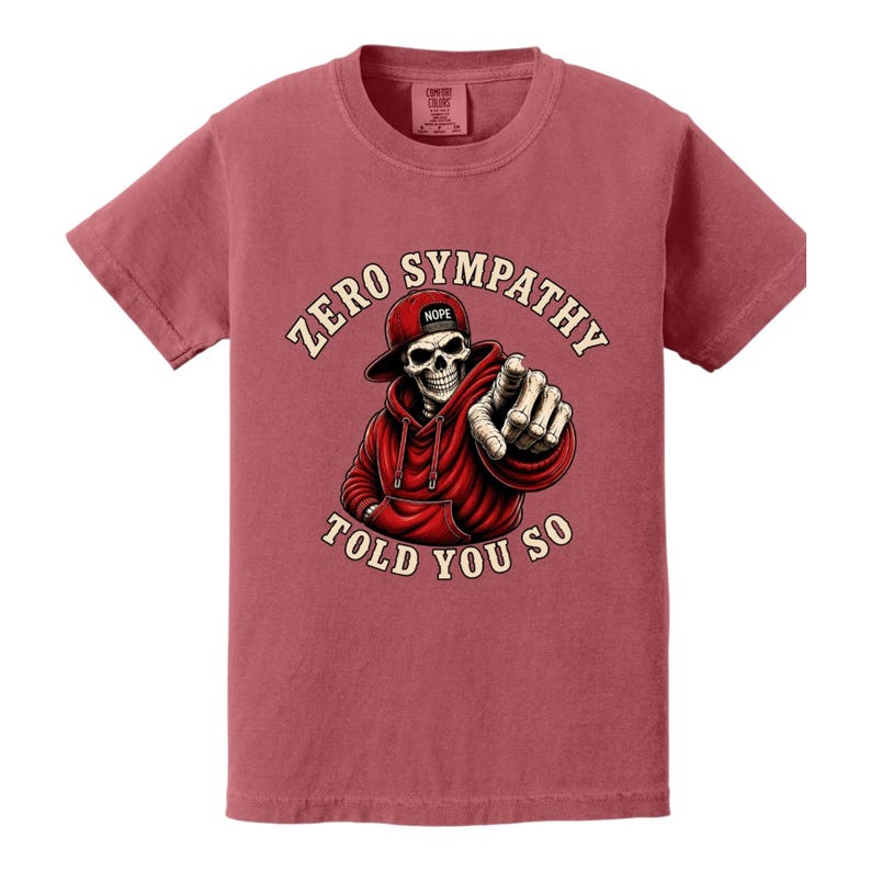 May include: A maroon t-shirt featuring a graphic of a skeleton wearing a red hoodie and hat, pointing. The text reads "ZERO SYMPATHY TOLD YOU SO". The hat has the word "NOPE" on it.