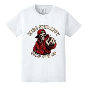 May include: White t-shirt featuring a graphic of a skeleton wearing a red hoodie and baseball cap. The skeleton is pointing with its index finger. Text on the shirt reads "ZERO SYMPATHY TOLD YOU SO."