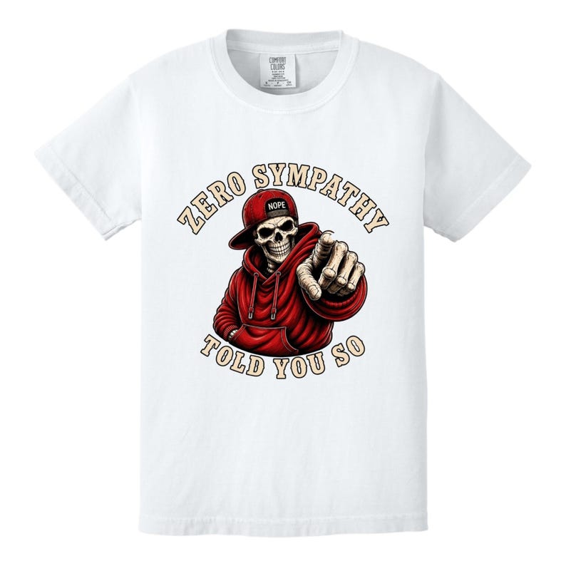 May include: White t-shirt featuring a graphic of a skeleton wearing a red hoodie and baseball cap. The skeleton is pointing with its index finger. Text on the shirt reads "ZERO SYMPATHY TOLD YOU SO."