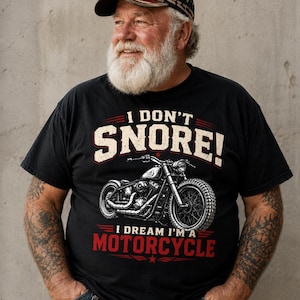 Pode incluir: Camiseta preta com o texto "I DON'T SNORE! I DREAM I'M A MOTORCYCLE" e um gráfico de motocicleta. A pessoa está usando um boné de beisebol com a bandeira dos EUA.