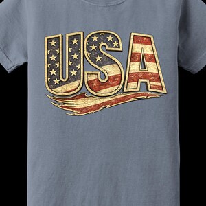 May include: A blue Comfort Colors t-shirt featuring the word "USA" in a distressed, patriotic design. The letters are filled with the American flag's stars and stripes, with a vintage, worn look.