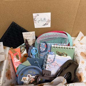 Puede incluir: Una caja de regalo de cart&oacute;n que contiene varios art&iacute;culos, incluyendo un estuche negro texturizado, una bolsa de malla verde menta, cuadernos florales, un plato azul, un tubo de crema de manos, una pinza para el cabello marr&oacute;n, un broche de mariposa y una pegatina con el texto Gabrielle's Gathering.