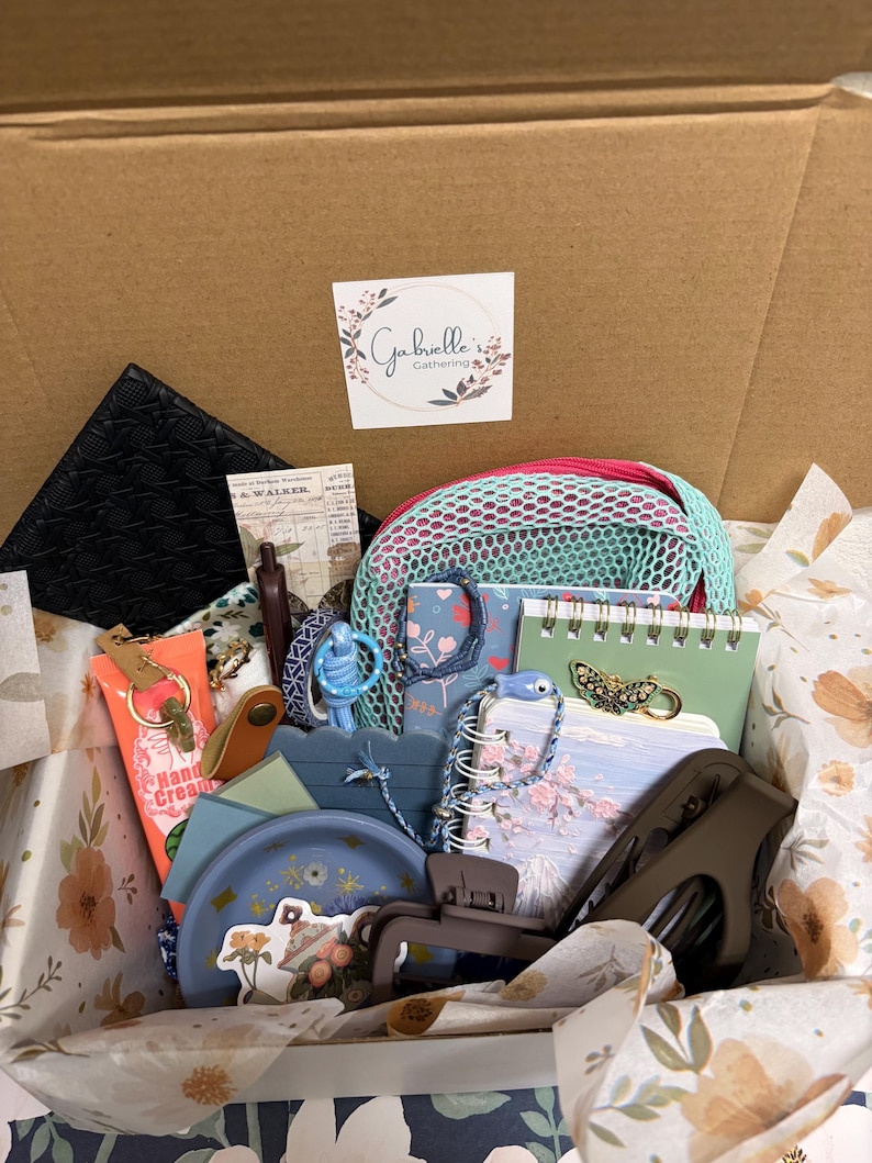 Puede incluir: Una caja de regalo de cart&oacute;n que contiene varios art&iacute;culos, incluyendo un estuche negro texturizado, una bolsa de malla verde menta, cuadernos florales, un plato azul, un tubo de crema de manos, una pinza para el cabello marr&oacute;n, un broche de mariposa y una pegatina con el texto Gabrielle's Gathering.