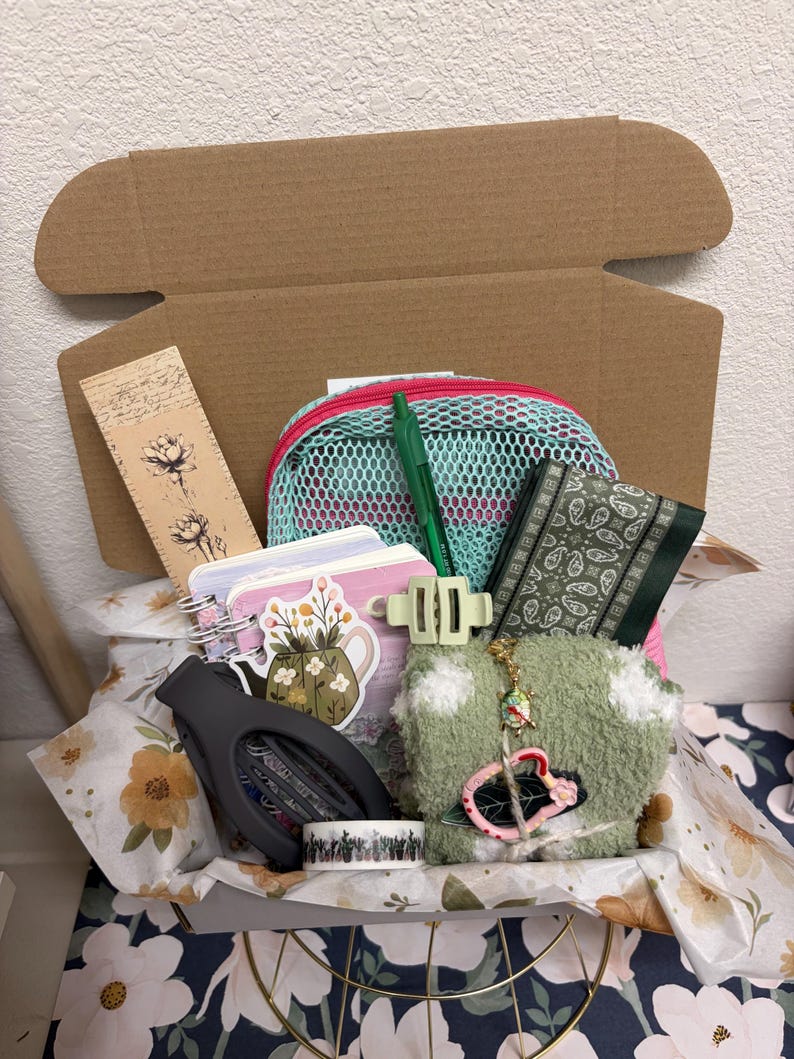 Puede incluir: Una caja de cart&oacute;n que contiene varios art&iacute;culos, incluyendo una bolsa de malla, un bol&iacute;grafo verde, un peque&ntilde;o cuaderno de espiral con una pegatina floral, una cartera verde oscuro con estampado paisley, calcetines verdes y un marcap&aacute;ginas.