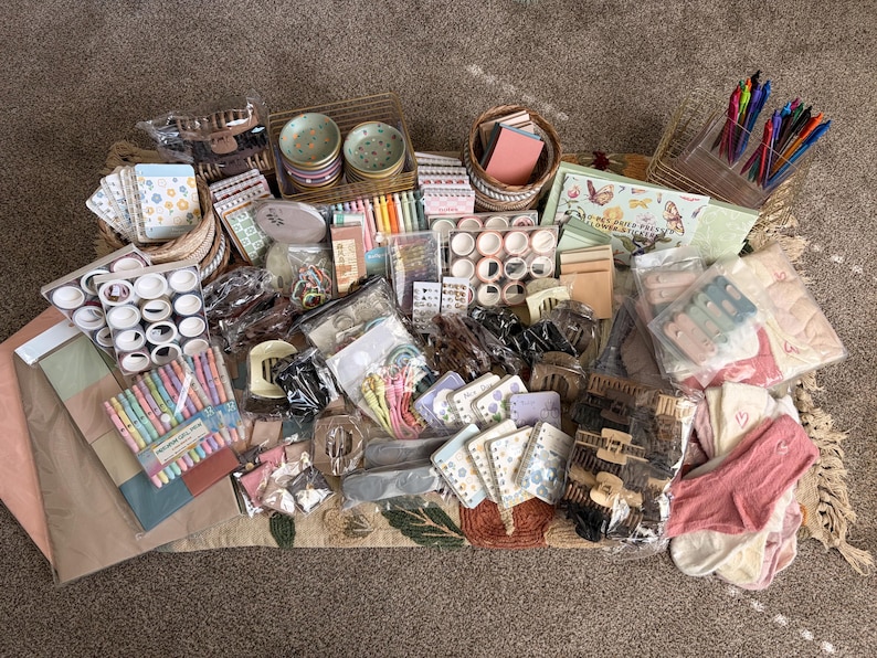 Puede incluir: Una colecci&oacute;n de art&iacute;culos de papeler&iacute;a y accesorios que incluye bol&iacute;grafos de colores, varios rollos de cinta washi, cuadernos peque&ntilde;os con motivos florales, pinzas para el cabello, calcetines y platos de cer&aacute;mica, todo en un gran surtido.